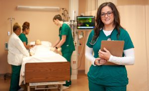 Female student dressed in green scrubs, holding a clipboard, and standing in front of 3 others in nursing simulation lab, who are working on a mannequin.