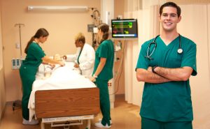 Male student dressed in green scrubs, smiling with arms folded and standing in front of 3 others in nursing simulation lab, who are working on a mannequin.